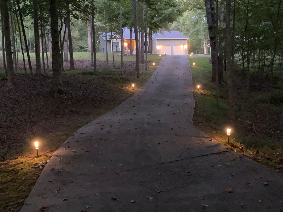 A well-lit driveway lined with soft glowing path lights leads toward a home surrounded by trees. The faint outline of a garage is visible, complemented by the warm light emanating from the house's windows, creating a welcoming entrance.