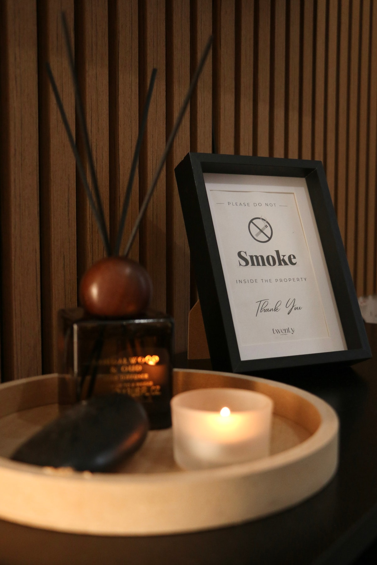 An elegant tabletop display features a framed sign reminding guests about the no smoking policy. A simple glass candle holder sits beside a round tray containing decorative elements such as a dark bottle and a wooden item, all set against a backdrop of warm wood paneling.
