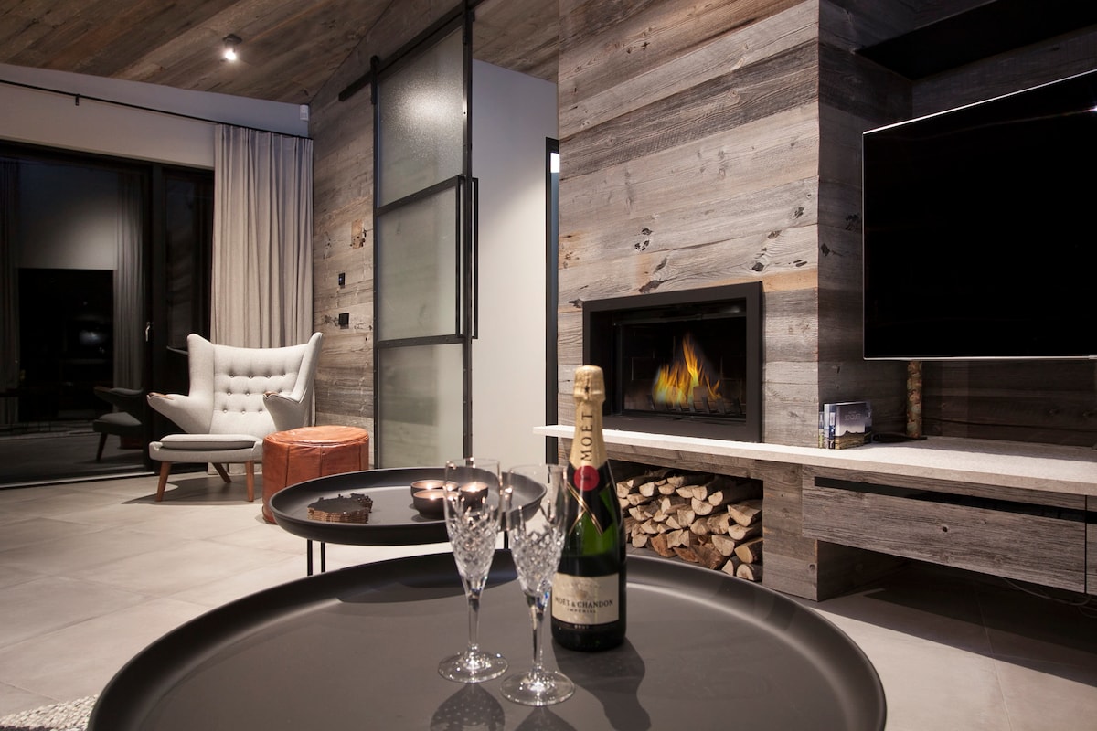 A modern living space is highlighted by a sleek stone fireplace and a flat-screen TV. A table is set with two crystal glasses and a bottle of champagne, while a cozy armchair and wooden accents enhance the composition.