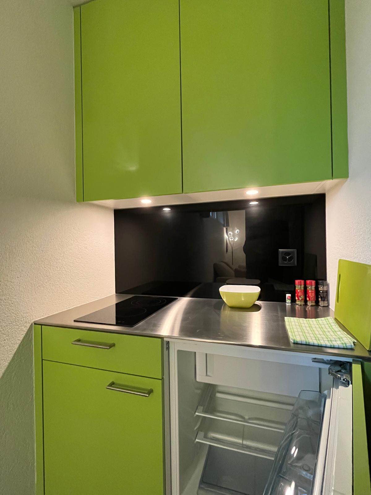 A compact kitchen area features green cabinetry contrasting with a sleek black backsplash. A stainless steel countertop includes a stovetop, and a small bowl sits atop it. An open refrigerator, displaying its empty interior, is also visible.