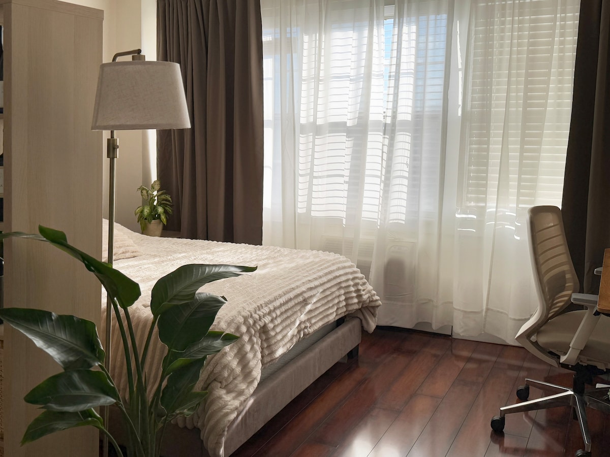 A cozy sleeping area features a spacious bed adorned with a light-colored quilt. Natural light filters through large windows, creating a warm ambiance. A green plant adds a touch of nature, while the polished wood flooring complements the room's contemporary design.