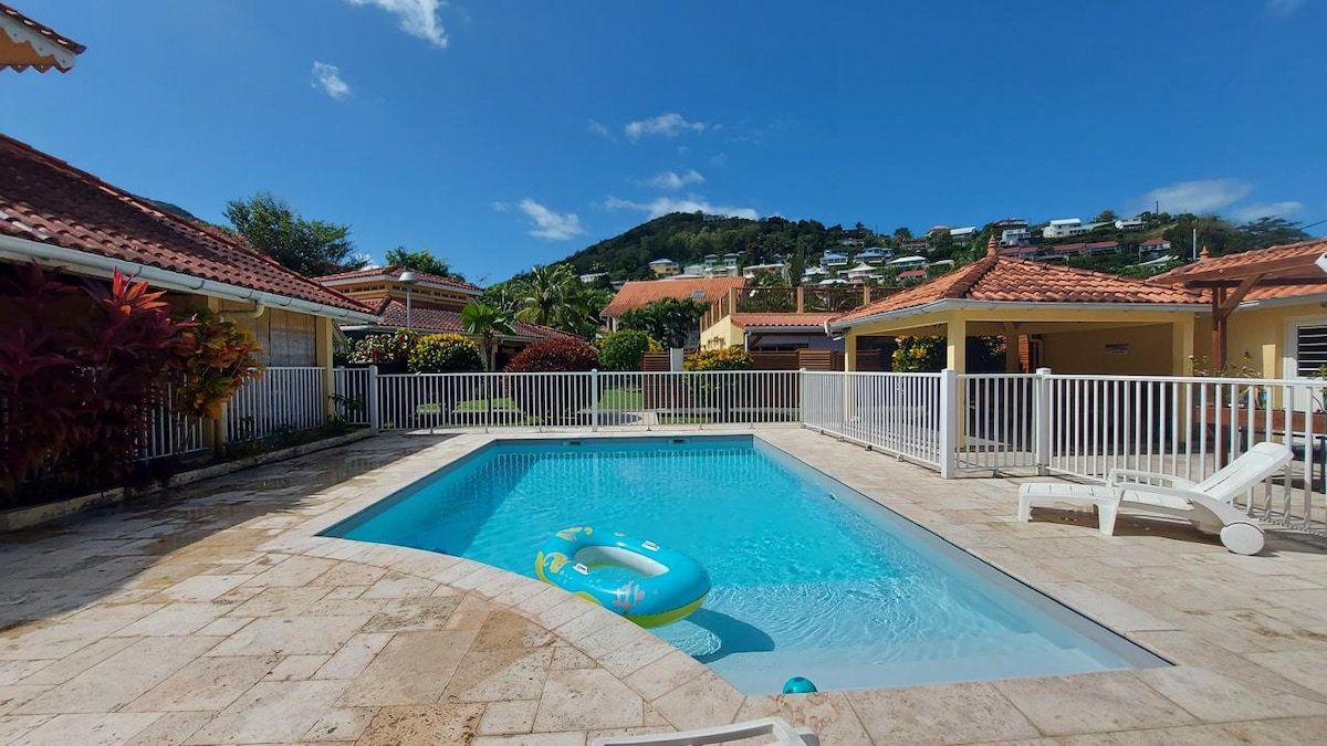A clear blue pool is surrounded by a marble deck and enclosed by a white safety fence. Tropical plants add color along the edges, with a sun lounger's white frame positioned nearby. A hillside with houses is visible in the background under a bright blue sky.