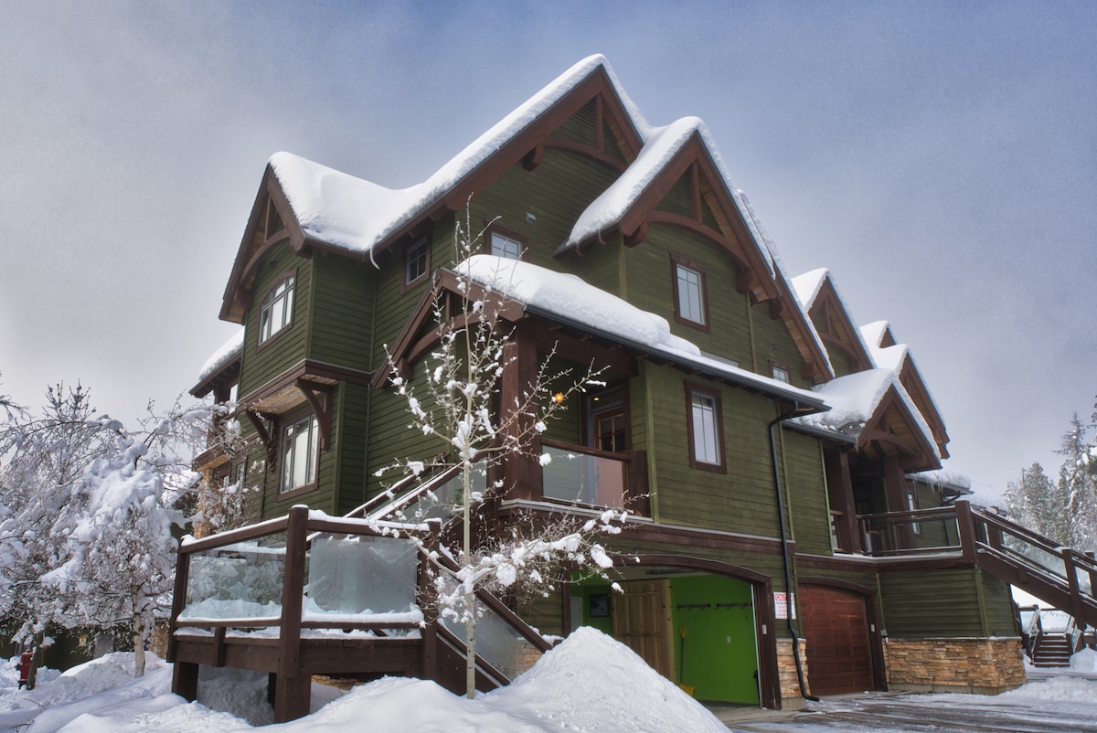The three-story green structure is surrounded by a snowy landscape, with a sloped roof covered in fresh snow. Large windows are visible, and wooden accents add warmth. A balcony with glass railing provides outdoor space, while a garage is partially seen in the background.