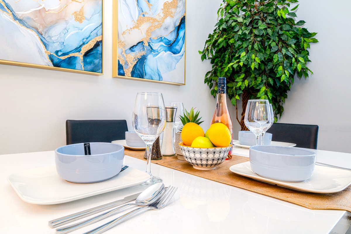 A modern dining table is set with four place settings, featuring light blue bowls, white plates, and polished silverware. A centerpiece of fresh fruit, including lemons and greenery, enhances the inviting atmosphere, while abstract artwork adorns the wall above.