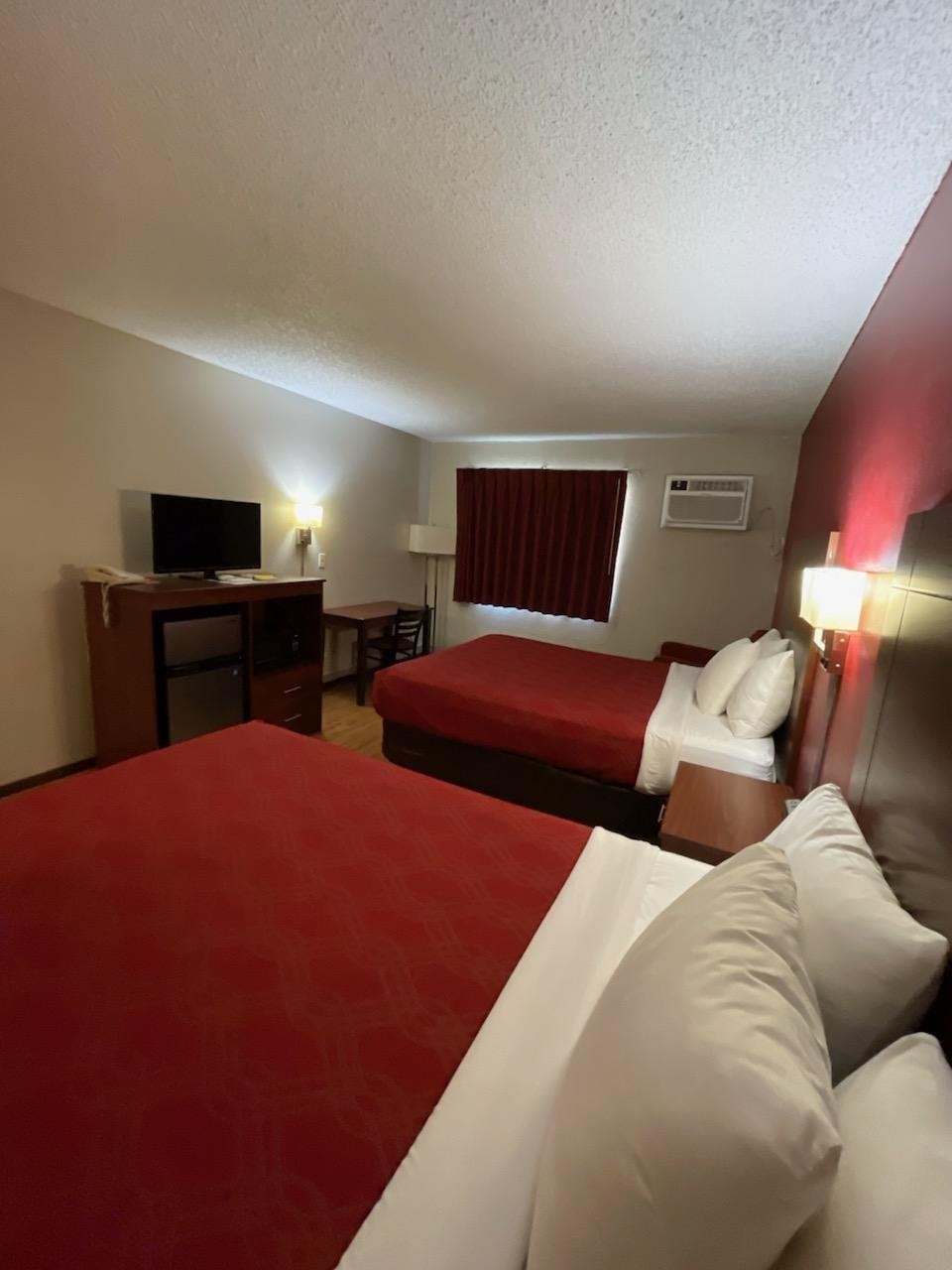 The guest room features two queen beds with red coverings, positioned against a textured wall. A functional workspace is visible, along with a television and a small refrigerator. Natural light enters through a window adorned with dark curtains, providing a comfortable and inviting ambiance.