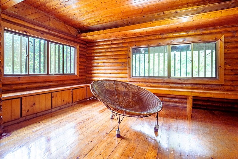 A spacious interior features wooden log walls and a polished wooden floor. Light streams through multiple large windows, enhancing the natural ambiance. A distinctive round chair is prominently placed in the center, complementing the inviting layout designed for relaxation.