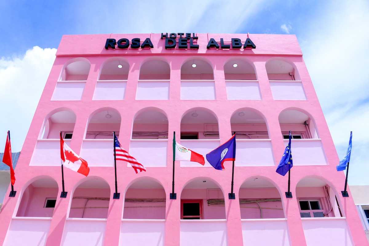 A vibrant pink hotel with multiple arches is featured, showcasing an inviting facade. Flags from various countries adorn the front, creating a diverse and welcoming atmosphere. A clear blue sky serves as the backdrop, enhancing the hotel's striking appearance.