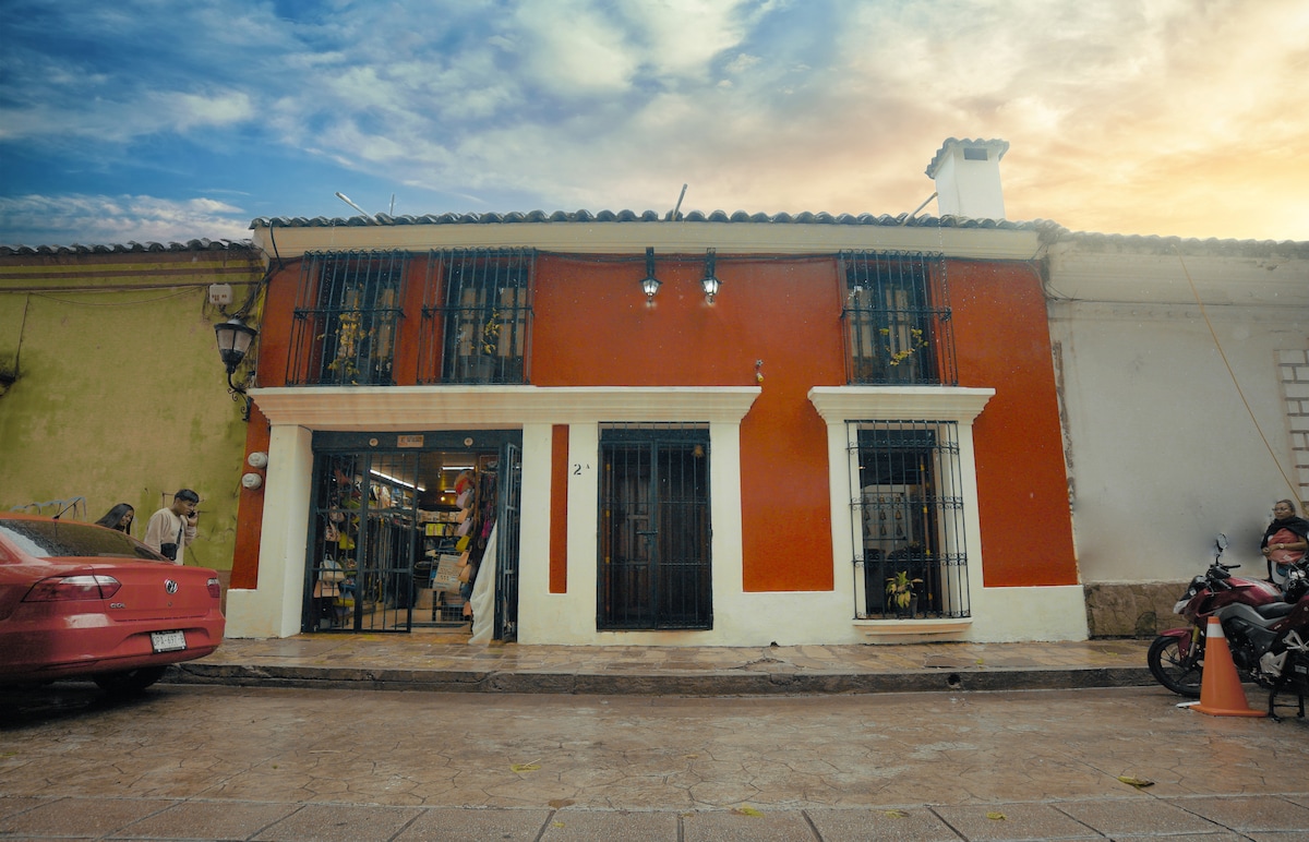A vibrant two-story building is presented with a combination of red and yellow facades. The main entrance is framed by decorative iron gates, while shops display local crafts within. A motorcycle is parked nearby, and a calm sky transitions from evening to twilight.