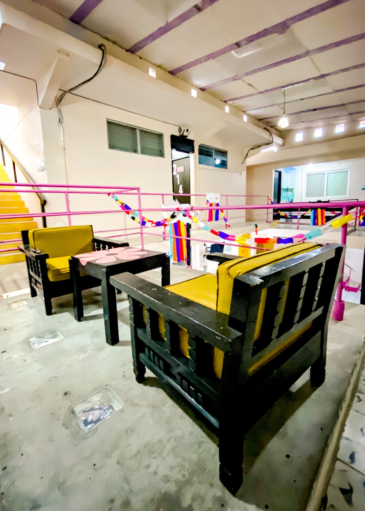 A casual seating area features two dark wood chairs with yellow cushions facing a small table. Brightly colored decorations are strung across the space, creating a cheerful atmosphere. The area is well-lit with overhead lights, and a staircase is visible in the background.