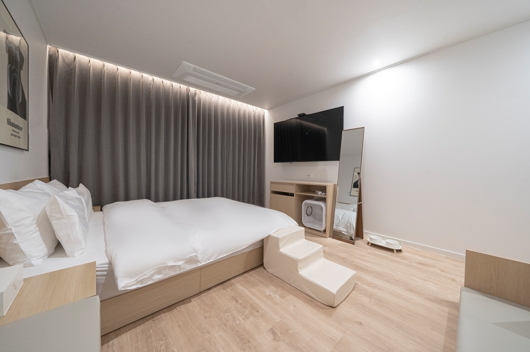 A bright and airy room features a spacious bed dressed in soft white bedding. A large flat-screen TV is mounted on the wall opposite. Natural light filters in through the floor-to-ceiling curtains, creating a calming atmosphere. A mirror and a small step are positioned nearby.