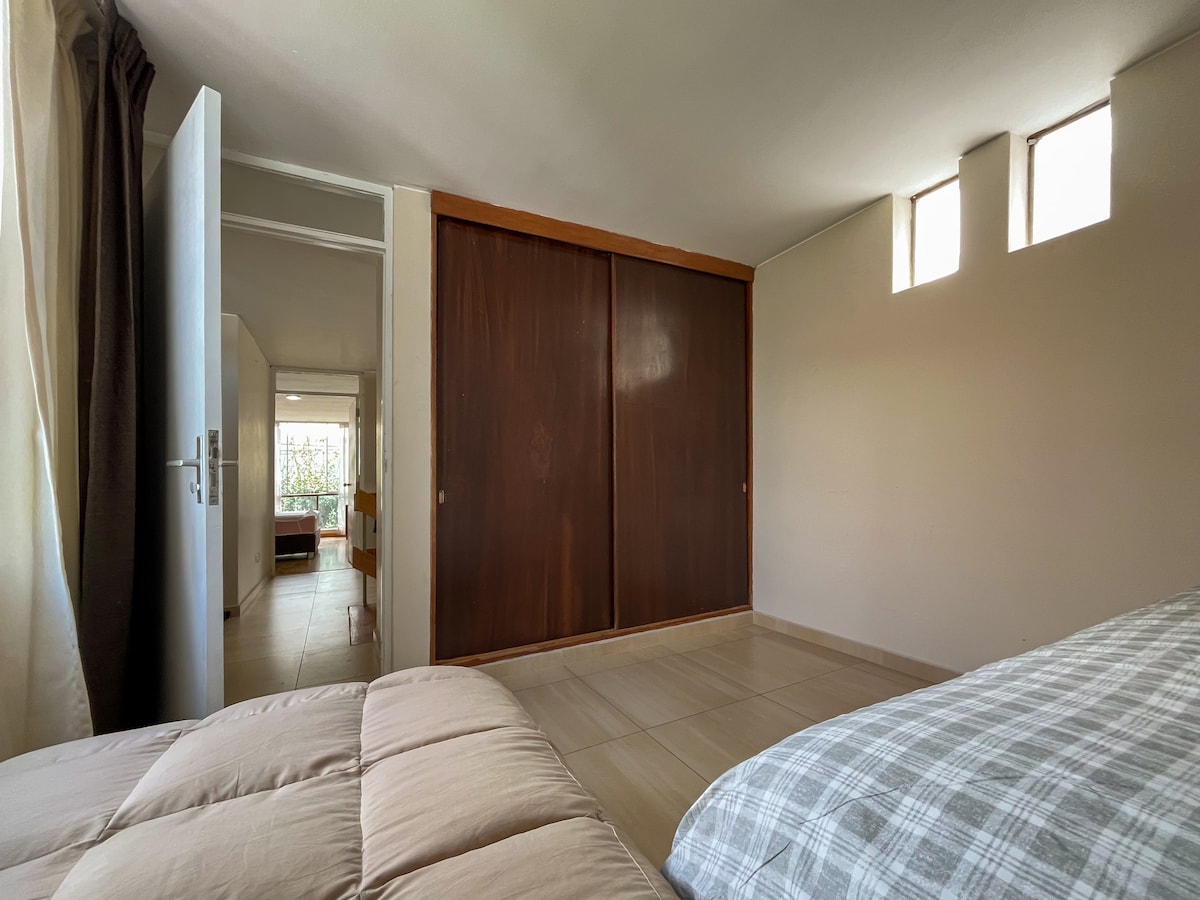 A spacious bedroom is presented, featuring a large built-in wardrobe with dark wooden doors. Natural light enters through three small windows, illuminating the tiled floor. A cozy bed with a neutral-colored blanket is visible, offering a comfortable resting space.