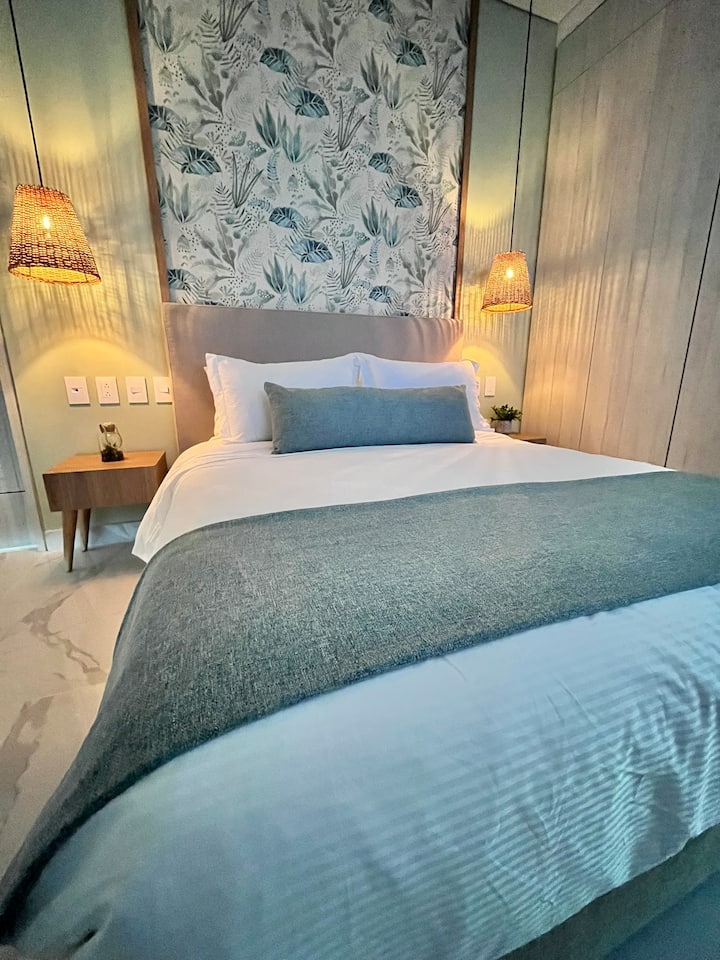 Cama doble con cojines decorativos, cabecera estampada y lámparas de lectura a ambos lados, creando un ambiente cálido y acogedor.