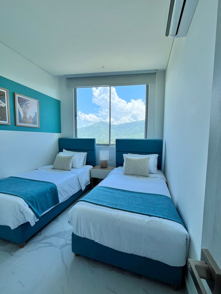 Dos camas individuales perfectamente organizadas, acompañadas de una gran ventana que ofrece una vista luminosa y despejada.