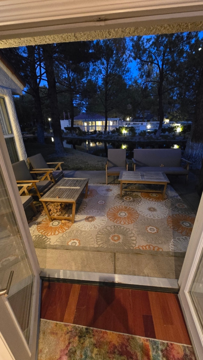 An outdoor patio area is shown illuminated by soft lighting in the evening. Comfortable seating is arranged around a patterned rug, with a view of the water in the background reflecting the glow from surrounding trees and structures.