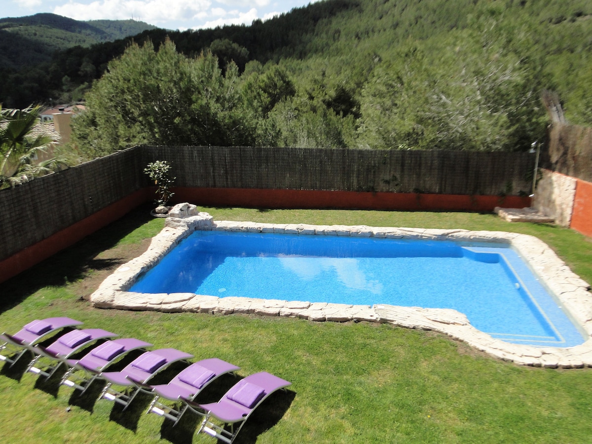 A private pool is visible within a well-maintained garden, surrounded by greenery and a low stone wall. Eight loungers are arranged on the grass nearby, providing ample space for relaxation. The pool's clear blue water reflects the natural light, offering a refreshing retreat.