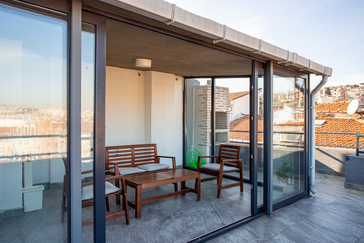 The shared rooftop terrace features a glass-enclosed seating area with a wooden table and benches. Natural light fills the space, offering views of the surrounding cityscape and rooftops. A small green plant completes the inviting atmosphere.