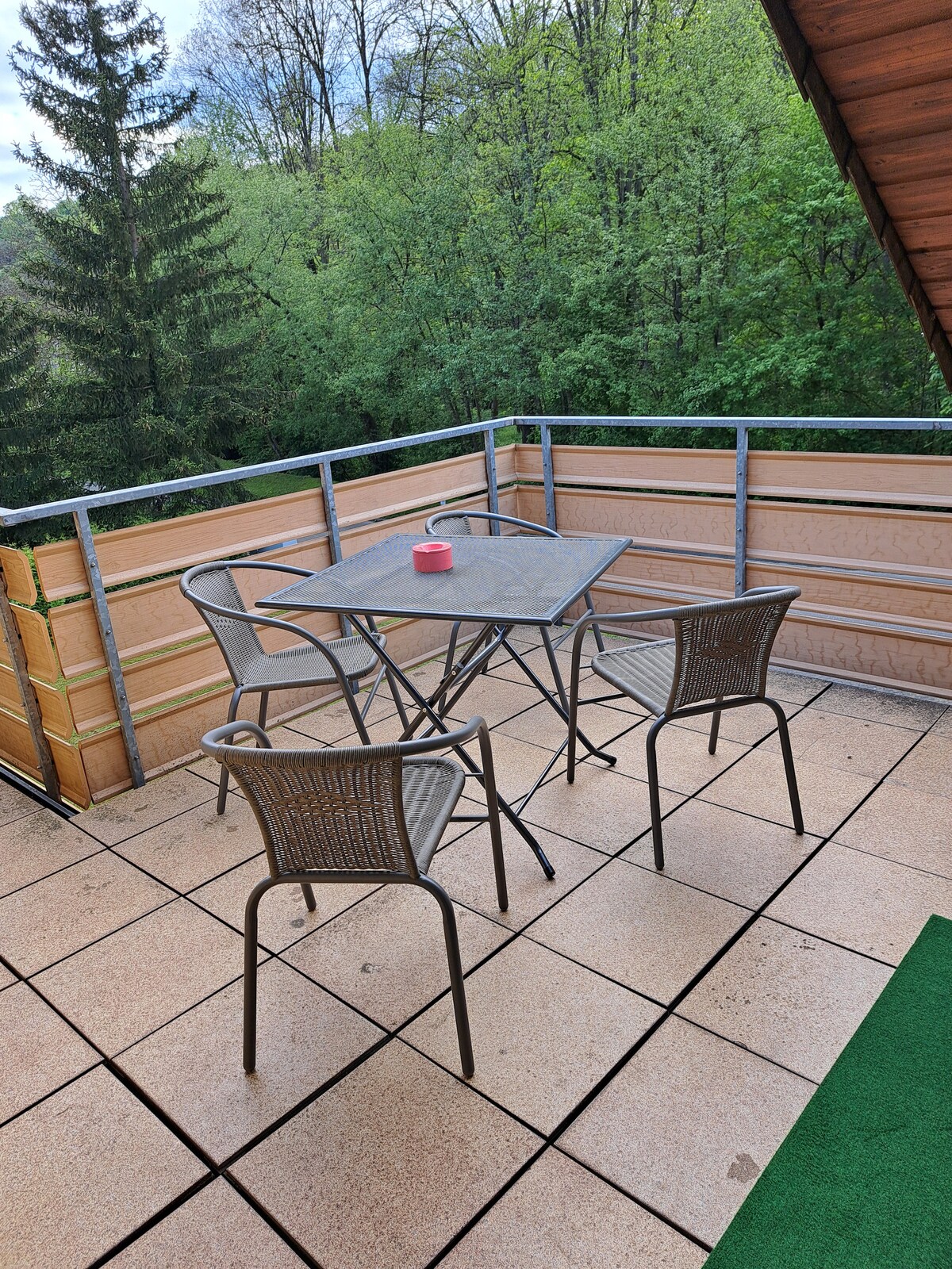 An outdoor balcony is furnished with a compact table surrounded by four woven chairs. The area features square tile flooring and is bordered by a railing. Lush greenery from surrounding trees provides a peaceful backdrop, enhancing the serene atmosphere.