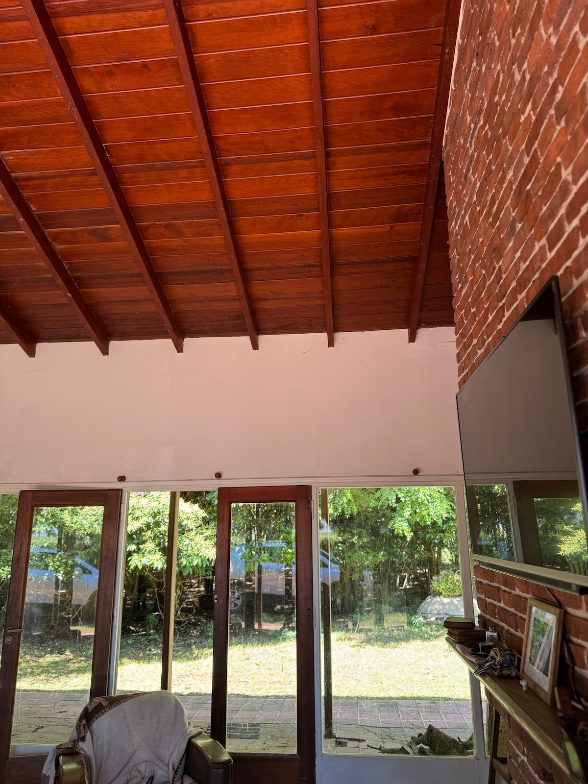 A cozy interior space features a high wooden ceiling with exposed beams. Large glass doors invite natural light and offer views of the outdoor greenery. A wall-mounted television is positioned above a wooden shelf decorated with small items, creating a warm and inviting atmosphere.