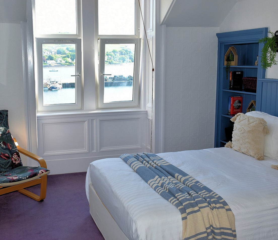 A serene bedroom features a spacious bed dressed in white linens, with a soft blue throw draped across it. A comfortable chair sits beside a bright window, offering views of a harbor. A bookshelf is positioned against the wall, adding a touch of color.
