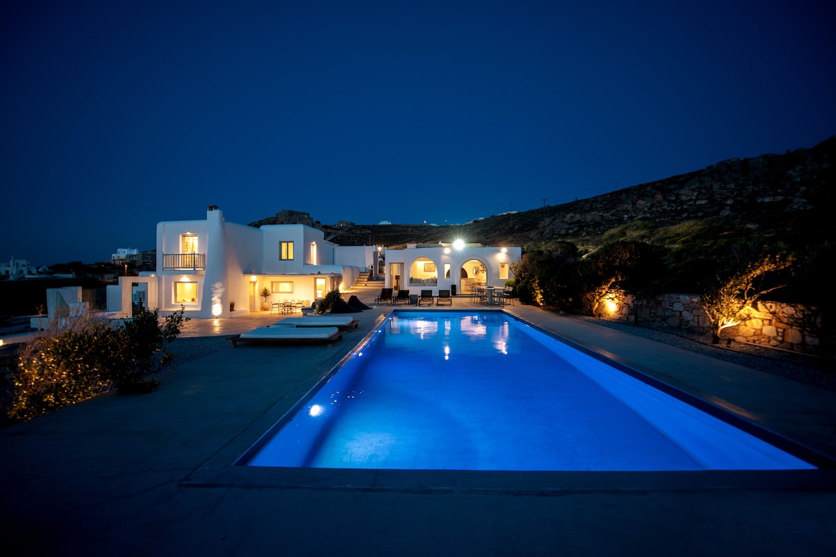 The villa is illuminated at night, showcasing a striking pool with a rich blue hue. Surrounding the pool, lounge chairs and well-maintained landscape accentuate the peaceful ambience. The two-story structure features elegant arches and large windows, creating a harmonious connection with the outdoor setting.