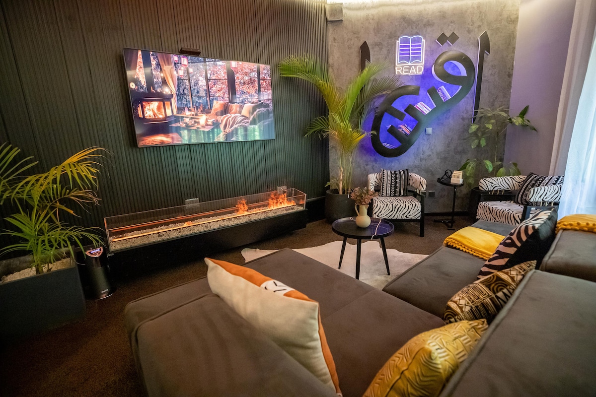 A modern lounge area is presented with a large wall-mounted television displaying a cozy scene. A low coffee table sits on a light area rug, surrounded by a sectional sofa adorned with colorful cushions. Indoor plants and a sleek wall design contribute to a relaxed ambiance.