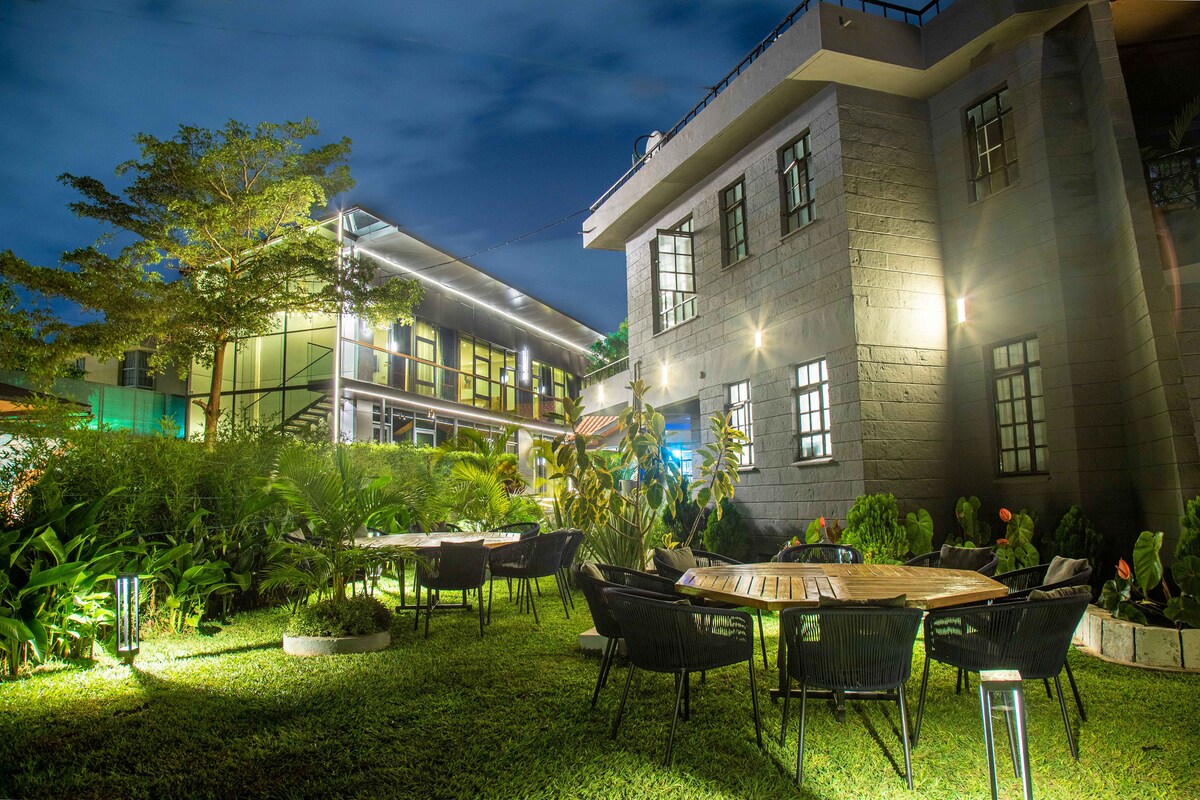 A captivating garden area is illuminated at night, featuring circular tables surrounded by black chairs. Lush greenery lines the space, while two modern structures are seen in the background, showcasing a mix of contemporary architecture and natural surroundings.