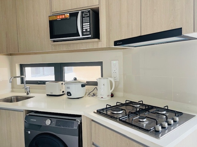 A modern kitchen features light wood cabinets and a clean countertop. A built-in sink is positioned next to a gas stove with five burners. Appliances, including a microwave and rice cooker, are arranged on the shelves, alongside a kettle, offering convenience for meal preparation.