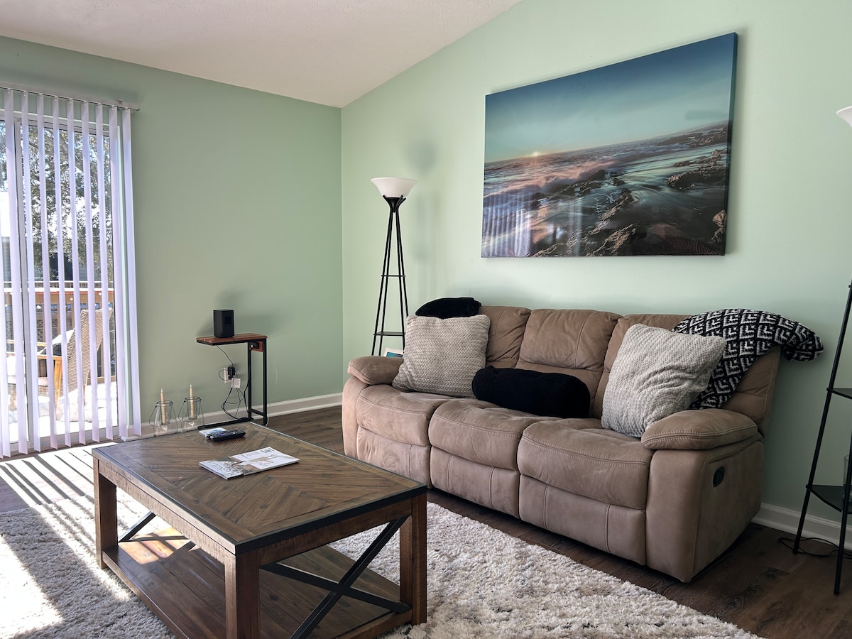 A spacious living room is showcased with a large, comfortable sofa adorned with decorative pillows. A wooden coffee table sits centrally, while a vibrant landscape painting hangs on the wall. Natural light filters through vertical blinds, illuminating the soft area rug beneath.