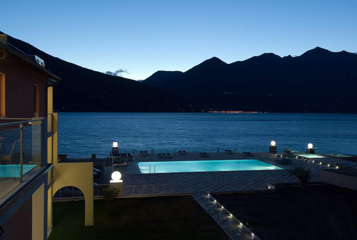 A serene evening view of the lake and mountains is presented, with soft lighting illuminating the outdoor pool area. The reflecting water and distant lights of the shoreline create a calm atmosphere, inviting relaxation and leisure.