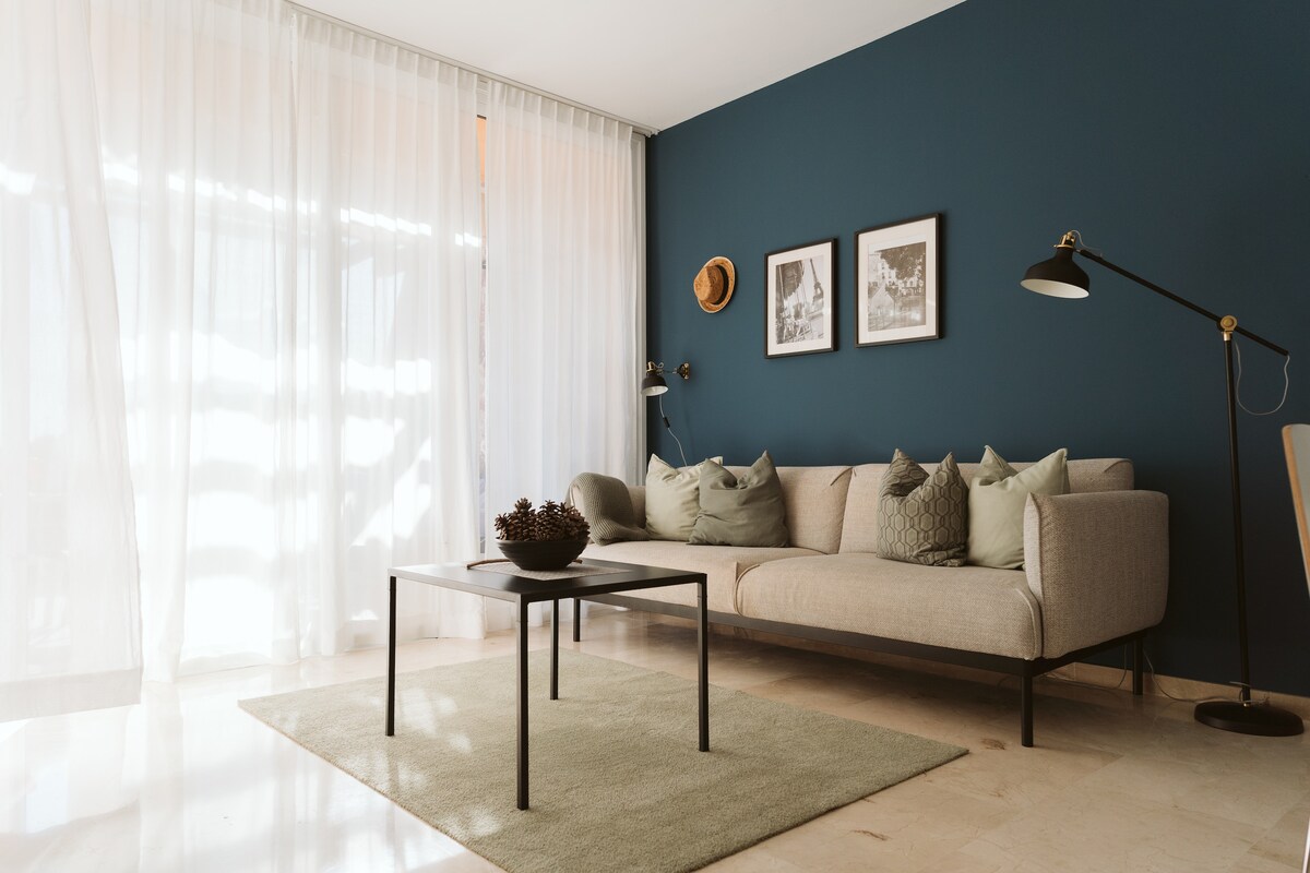 A light-filled living area features a beige sofa adorned with decorative pillows, positioned beside a small dark table. Large windows with sheer curtains allow natural light to brighten the space, while two framed artworks are displayed on the dark blue wall, enhancing the modern decor.