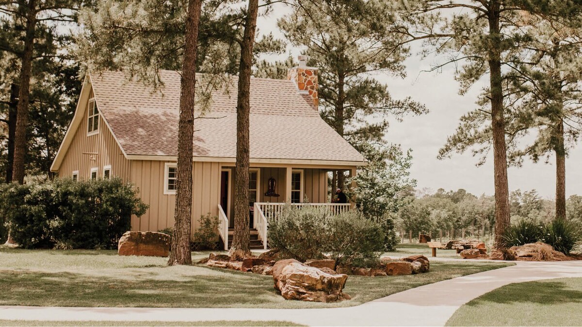A charming two-story cottage is nestled among towering pine trees, featuring a welcoming front porch and a light brown exterior. Surrounding rock formations add to the natural landscaping, while a paved path leads to the entrance.