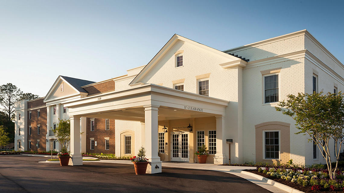 The resort's exterior features a welcoming entrance framed by large columns and a bright white façade. Lush landscaping with colorful flower beds enhances the curb appeal, while a smooth driveway leads to the entrance, creating a pleasant approach for guests.
