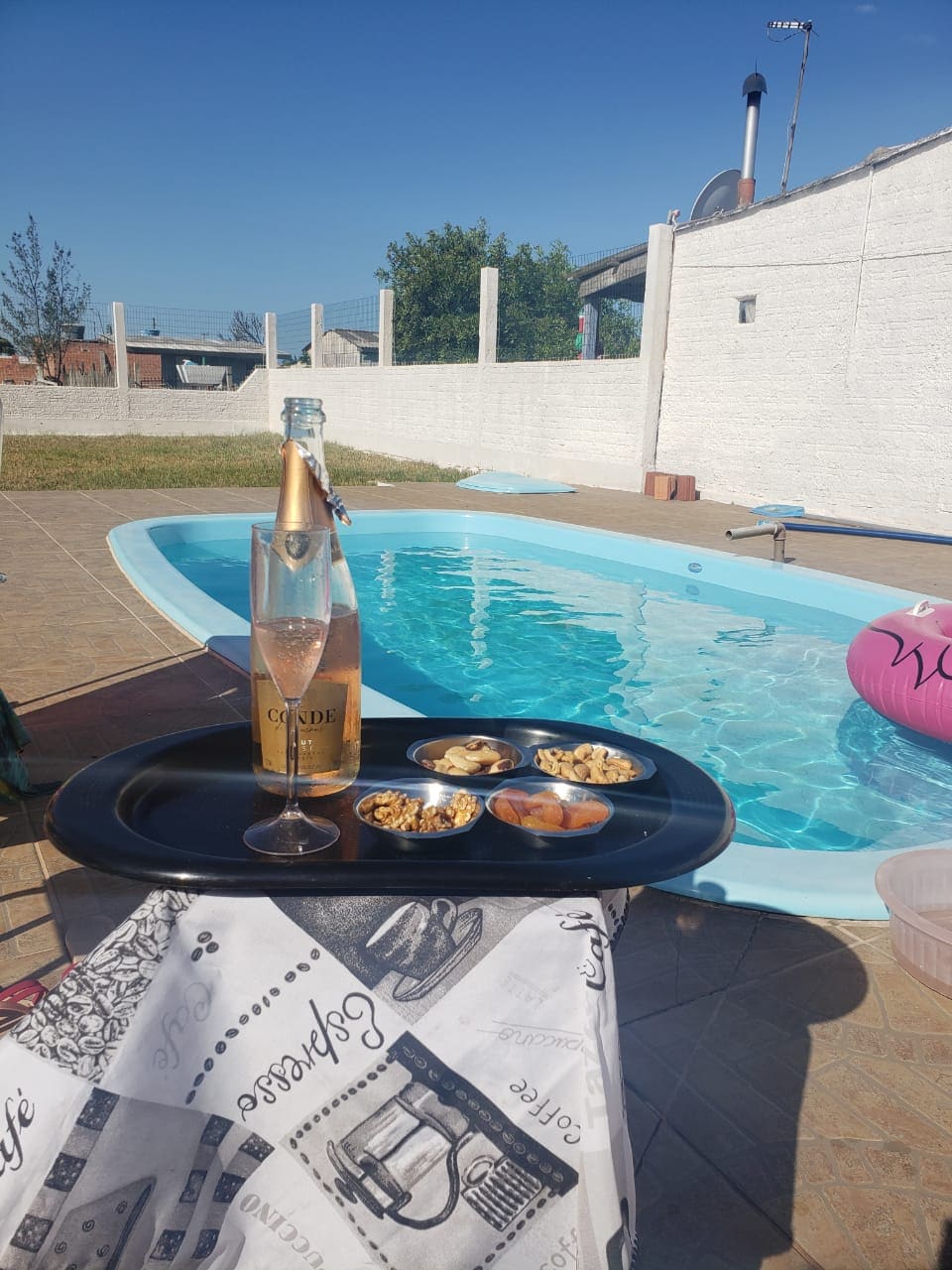 A sunlit patio area features a refreshing pool with clear water, surrounded by white walls. A tray with snacks and a champagne bottle sits in the foreground, inviting relaxation. Bright blue skies contribute to a serene outdoor setting.