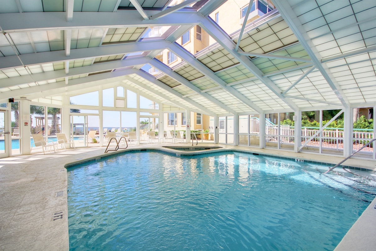 An indoor pool area is surrounded by large glass panels, allowing natural light to illuminate the space. The pool features a smooth, clear surface and is complemented by several lounge chairs arranged nearby. Views of the outdoor pool and surrounding greenery are visible through the transparent walls.