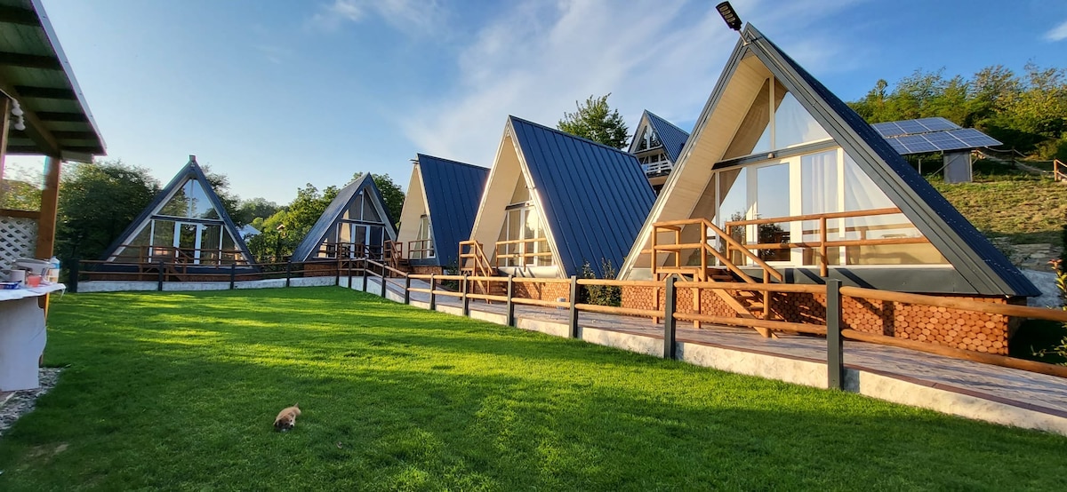 A cluster of modern A-frame cabins is set against a lush green landscape. Each cabin features wooden decks and large windows, allowing natural light to flood the interiors. Solar panels are visible on the hillside, emphasizing a commitment to sustainability.