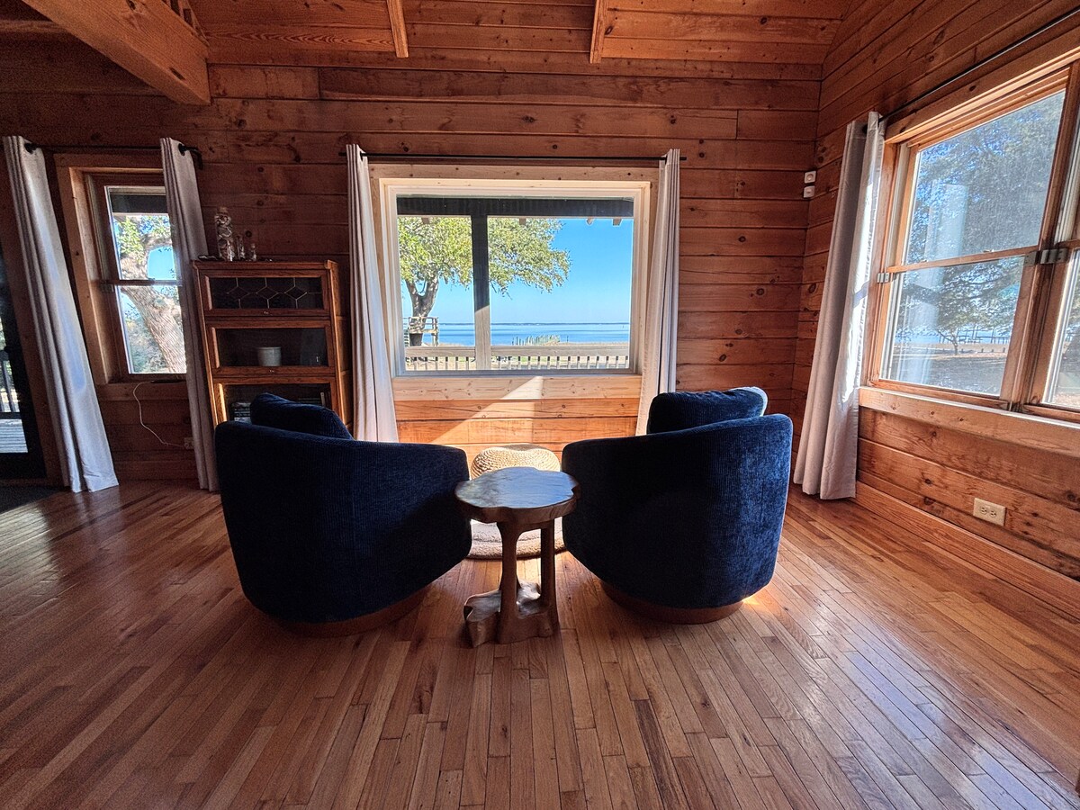 Two plush navy blue chairs are positioned around a small wooden table, set on polished hardwood flooring. Large windows allow natural light to illuminate the space, providing views of the expansive water scenery outside, framed by trees.