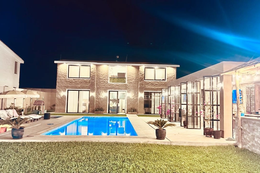 The open-concept house features sleek modern architecture, illuminated by soft lighting against a night sky. A clear blue pool is centrally positioned, surrounded by manicured grass and lounging areas. Expansive windows reflect the outdoor environment, creating a harmonious connection between indoor and outdoor spaces.
