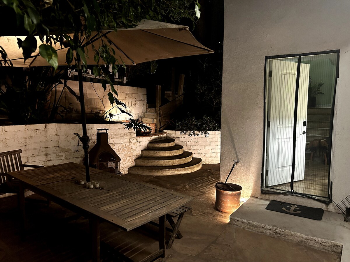 A private patio area is featured, showcasing a wooden table surrounded by benches. A large umbrella provides shade, while a decorative fire pit sits nearby. Curved stone steps lead to a white door, with soft lighting illuminating the pathway and surrounding foliage.