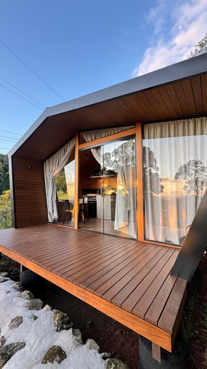 The exterior of the cabin showcases a modern design with a sloped roof and wooden siding. Large glass doors lead to a deck, framed by light curtains that softly filter the sunlight. Lush greenery and a clear sky are visible in the background.