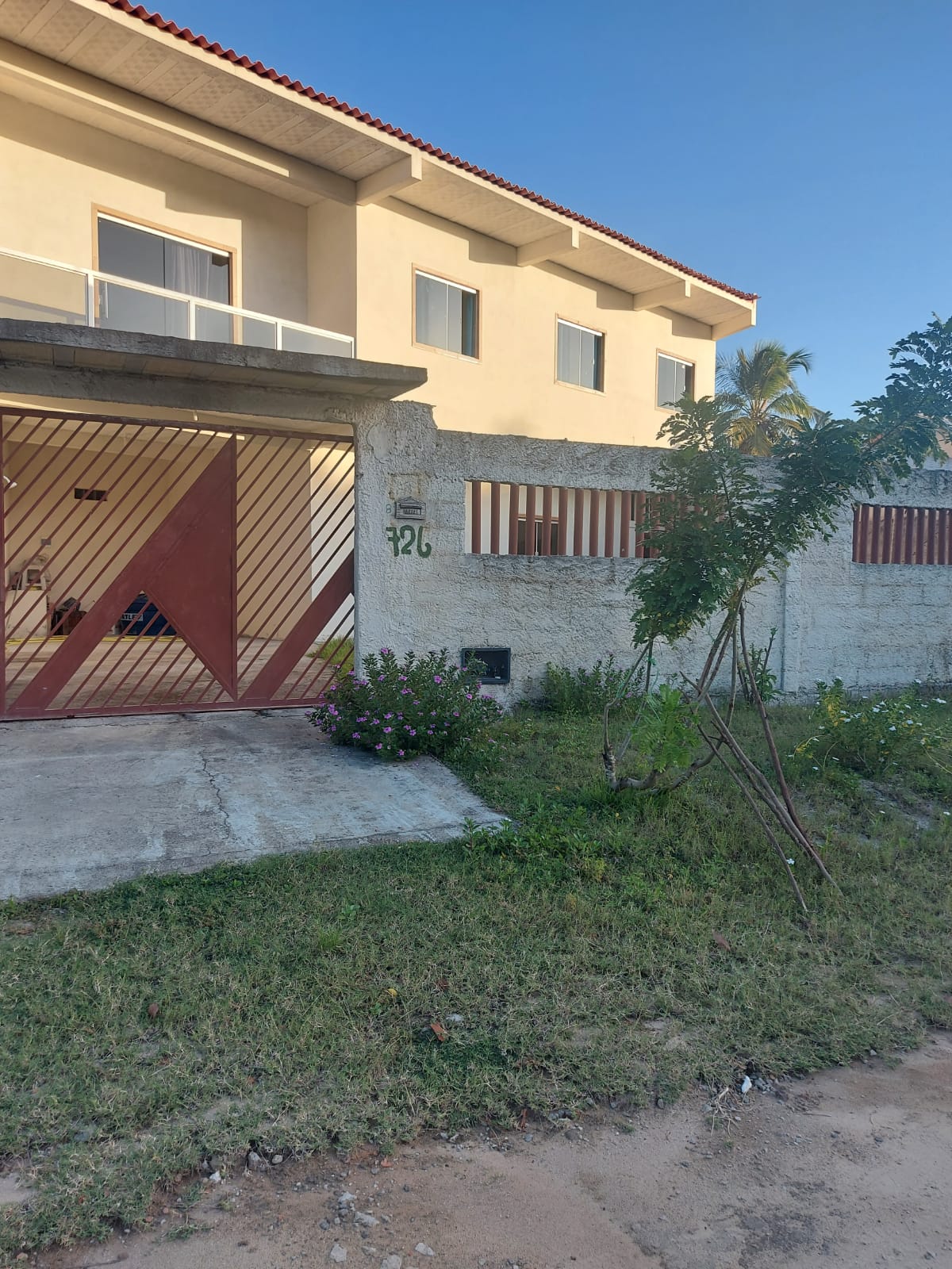 The exterior of a two-story house is shown, characterized by a combination of white walls and a red entrance gate. Lush green grass and small plants surround the entryway, providing a touch of nature. Clear blue skies create a serene backdrop.