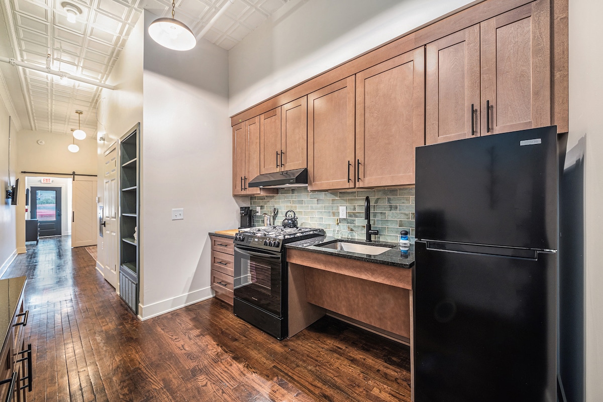 The kitchen area showcases modern cabinetry with a sleek black refrigerator and stove. Light brown cabinets complement the light-colored backsplash. The space features an open layout, leading to a hallway with various sources of natural light.