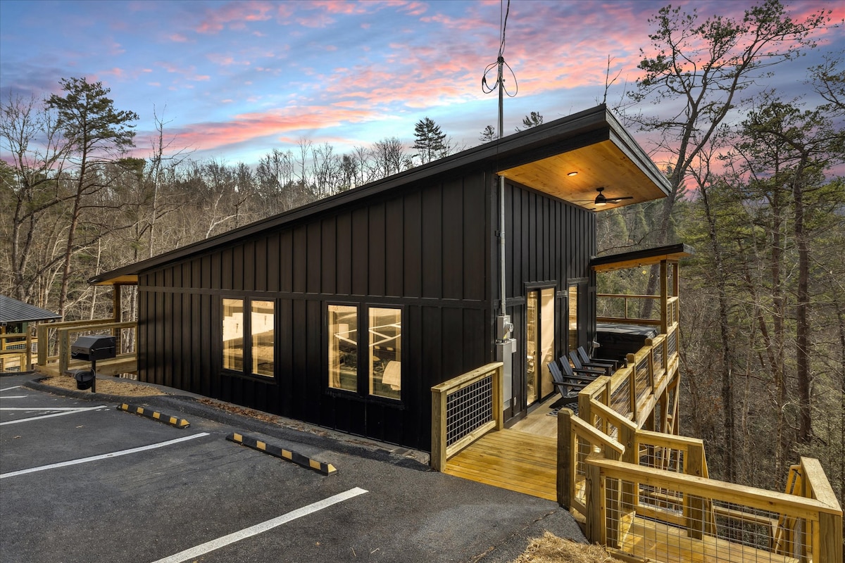 A modern treehouse cabin is nestled among the trees, featuring a dark wood exterior and large windows that provide natural light. The deck extends outward from the structure, with comfortable seating and a scenic view of the surrounding wooded area. Parking spaces are visible in the foreground.