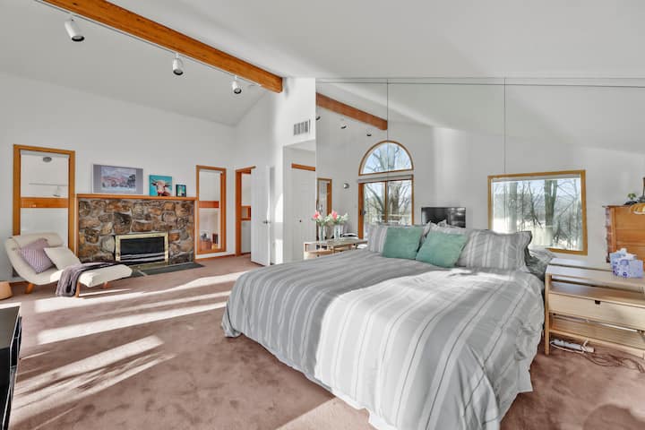 זווית נוספת של חדר שינה 3 עם קיר מראה, קמין אבן, פינת ישיבה ותקרות גבוהות.