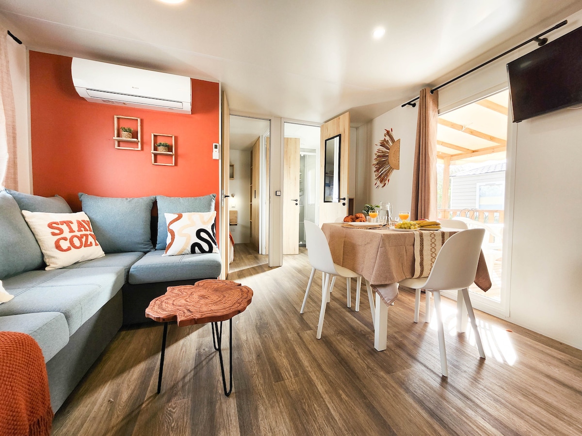 The interior space is shown, featuring a cozy seating area with a sectional sofa adorned with decorative pillows. A table with white chairs is set, while light floods through large glass doors that lead to an outdoor terrace, enhancing the open feel.