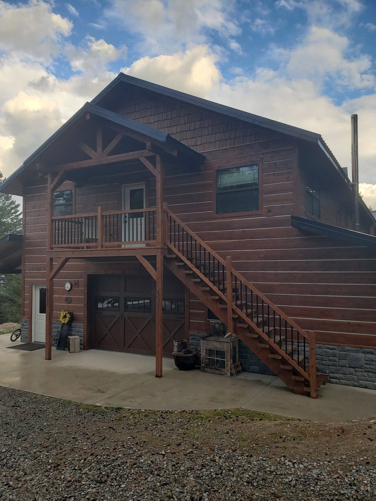 A rustic wooden cabin structure features a large porch with an overhang, supported by sturdy beams. A set of steps leads to the upper entrance, while a garage door is visible on the lower level. Natural surroundings complement the cabin's charm.