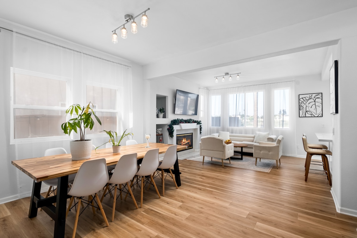 A bright dining and living area is shown, featuring a long wooden table surrounded by sleek chairs. Large windows allow ample natural light, highlighting the minimalist decor. A cozy seating arrangement with a fireplace is visible on one side, contributing to the inviting atmosphere.