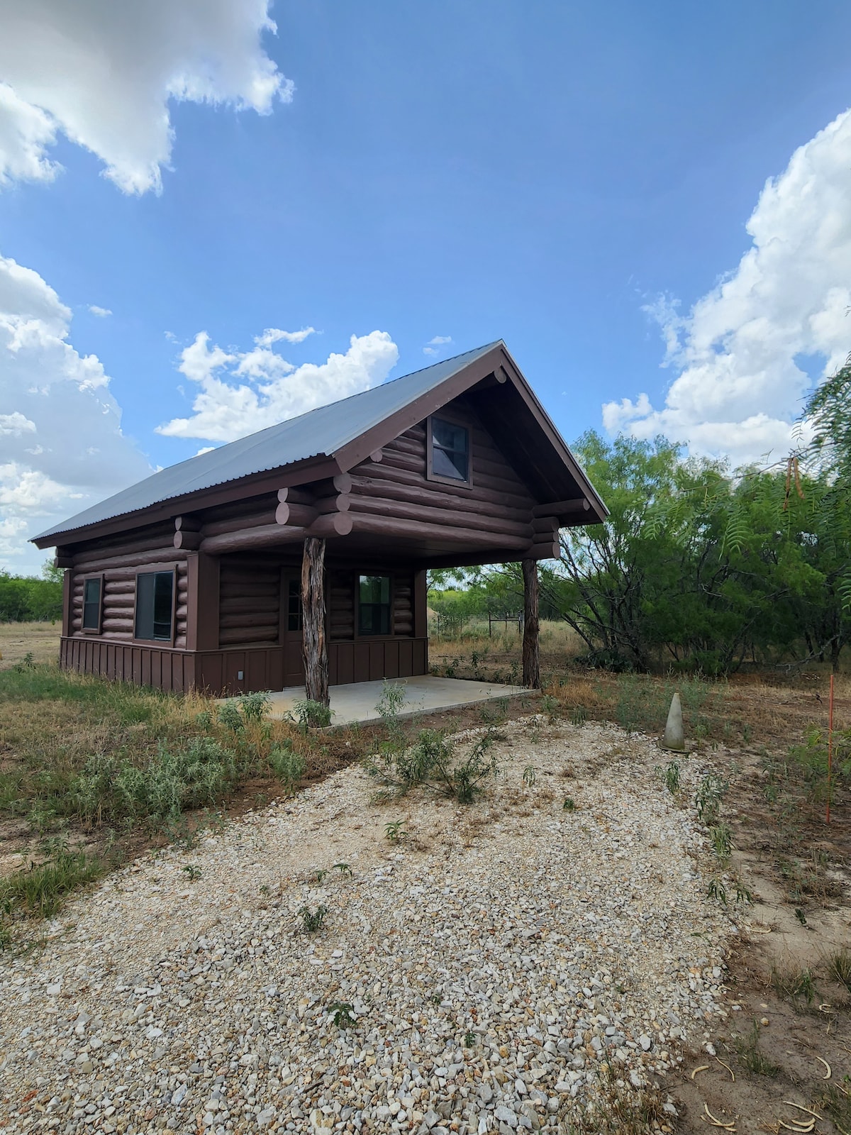 A log cabin is set against a blue sky with white clouds. The porch features wooden posts and an accessible entryway. Surrounding vegetation includes sparse shrubbery and grassy areas, indicating the cabin's rural setting.