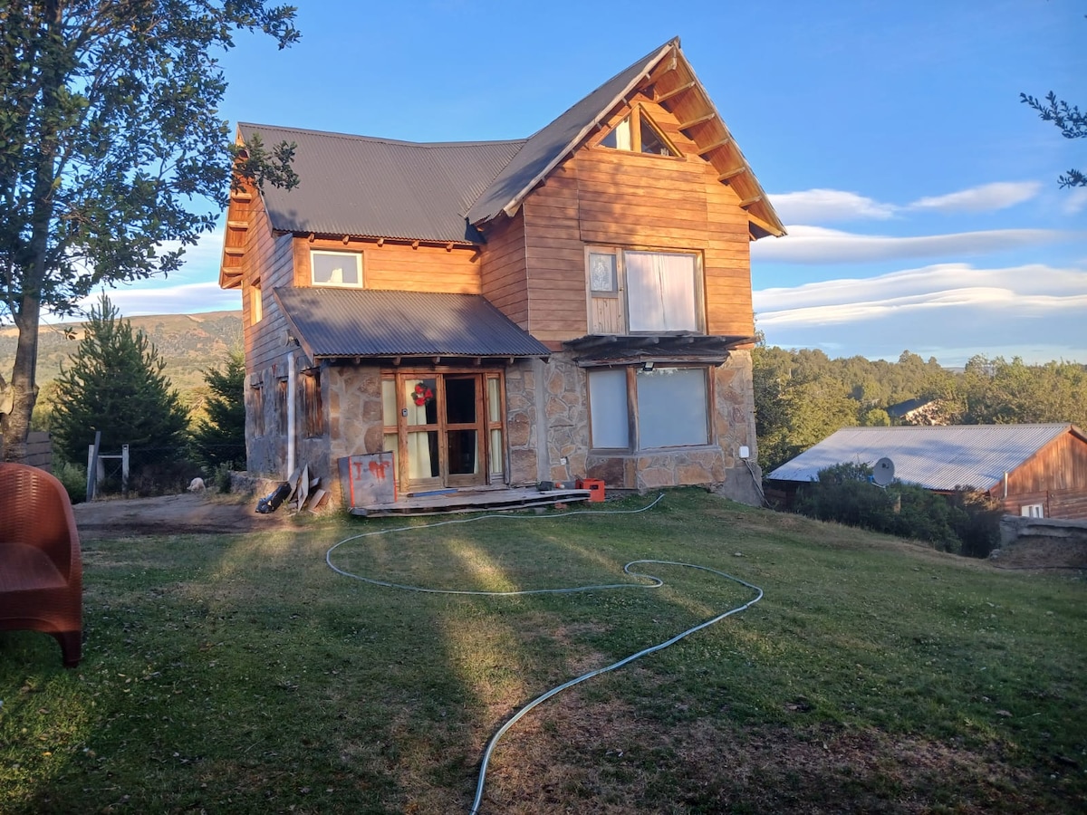 The two-story cabin features a wooden façade with large windows allowing natural light to fill the interior. A spacious lawn surrounds the structure, with a view of the natural landscape. A cozy outdoor seating area is visible in front, offering a welcoming atmosphere.