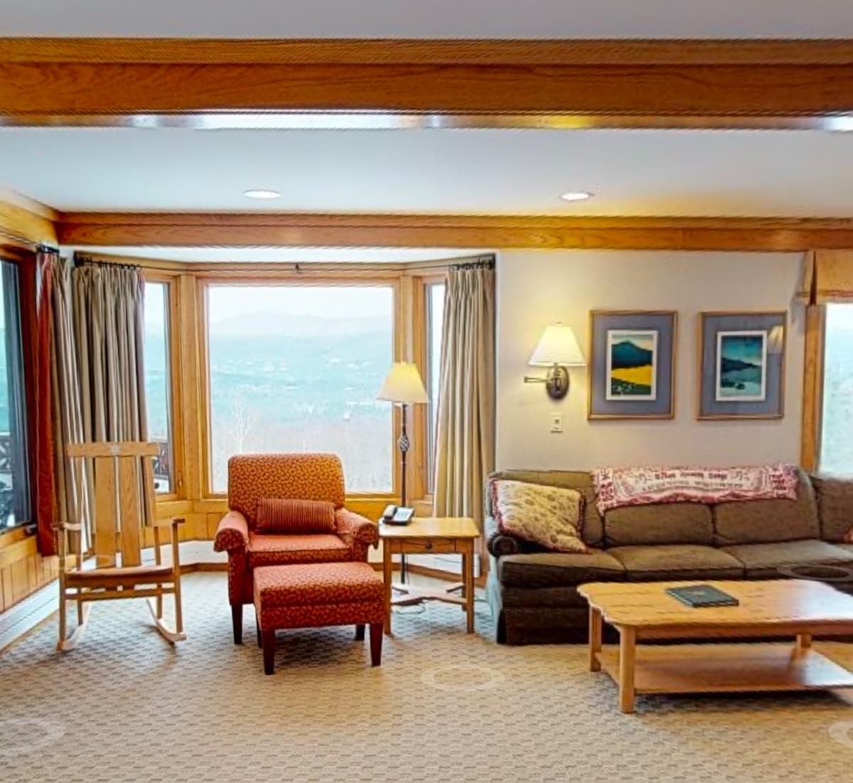 A spacious living area features a comfortable gray sofa, accented by a patterned armchair and natural wood tables. Large windows allow ample light and showcase scenic views. Soft curtains frame the windows, and artwork adorns the walls, adding a touch of personality to the space.