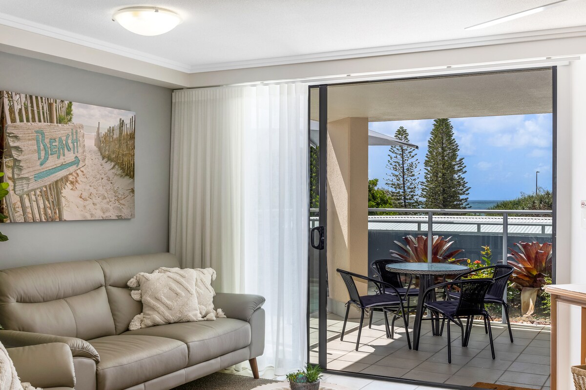 An open-plan lounge area features a light leather sofa and a beach-themed canvas artwork on the wall. Large glass doors provide a view of the patio with outdoor seating and glimpses of the ocean and surrounding greenery.